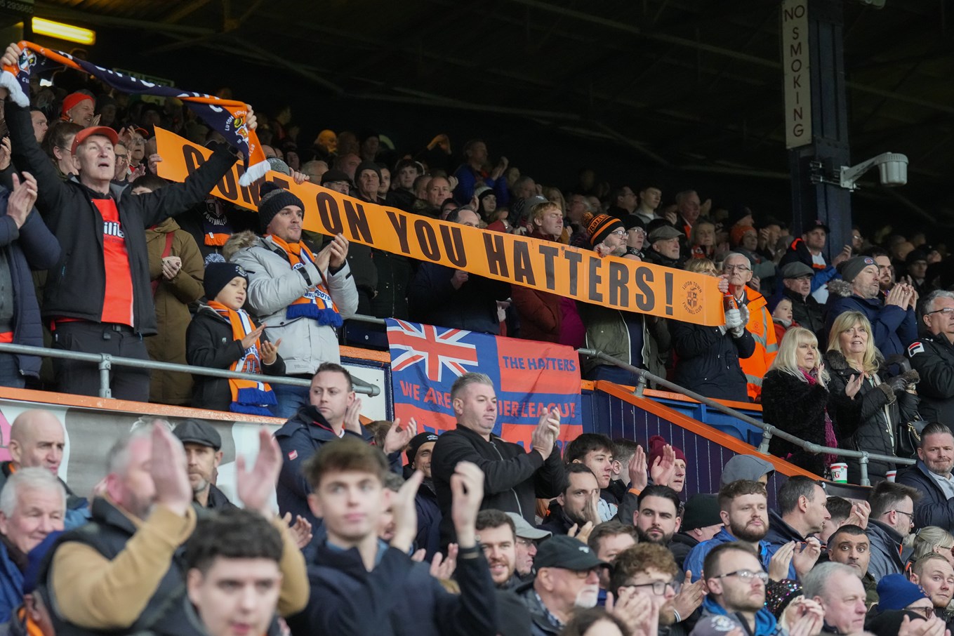 Luton Town fans in the Main Stand clapping with a banner raised which says 'Come on you Hatters!'.